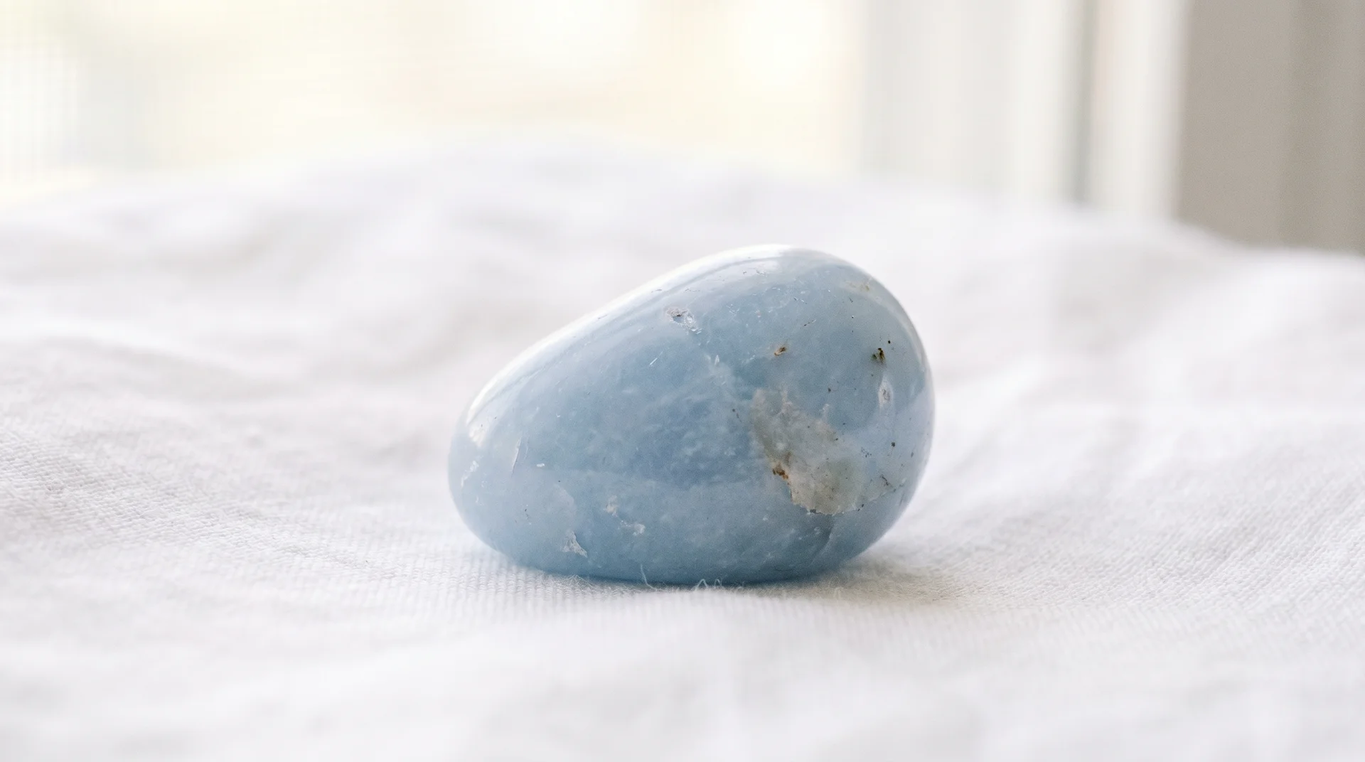 A close-up shot of a smooth, polished pale blue angelite stone resting on a textured white fabric surface.