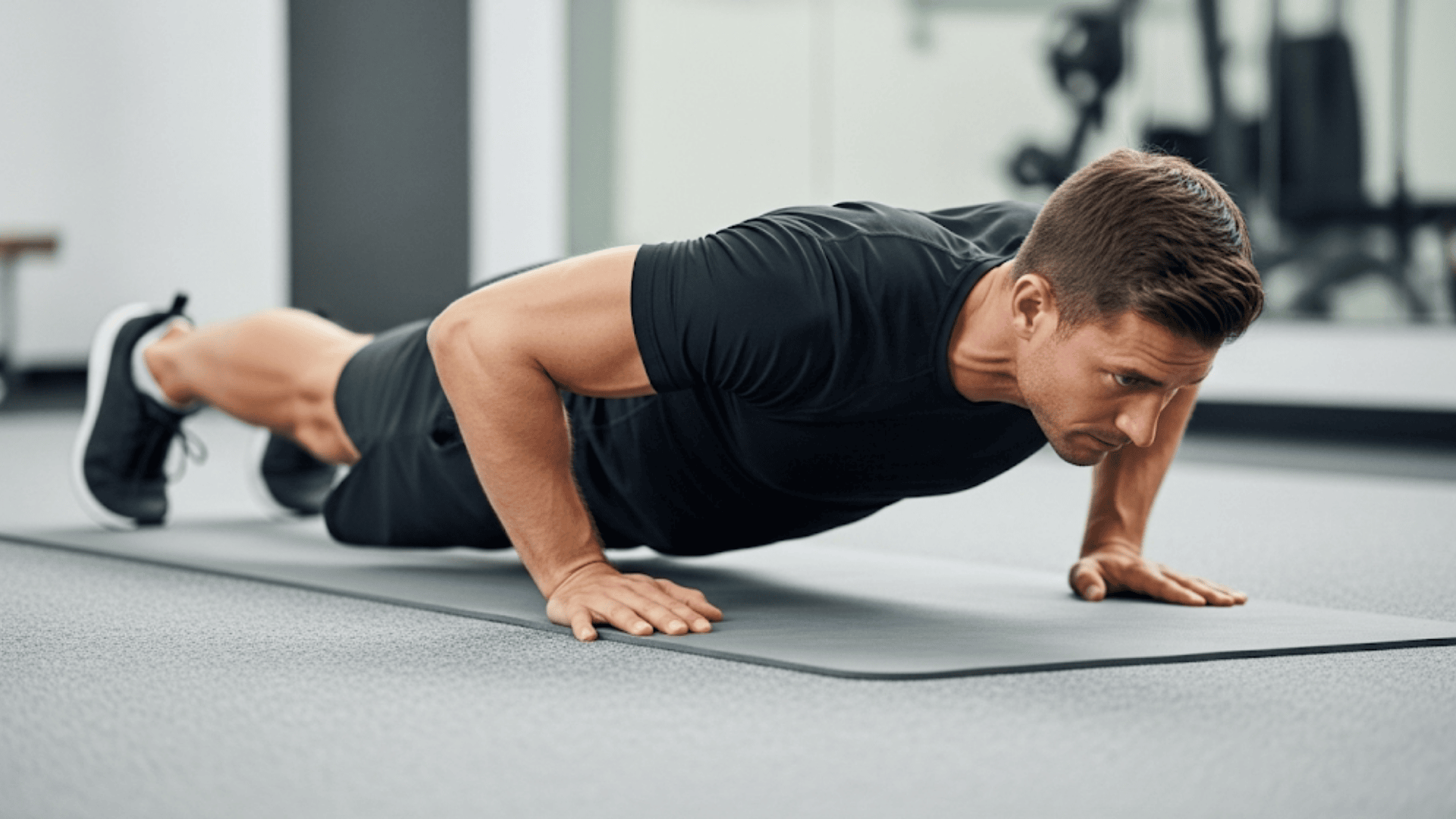 A man performs a push-up with a straight body, hands slightly wider than shoulders, elbows bent, and core engaged on a gym mat.