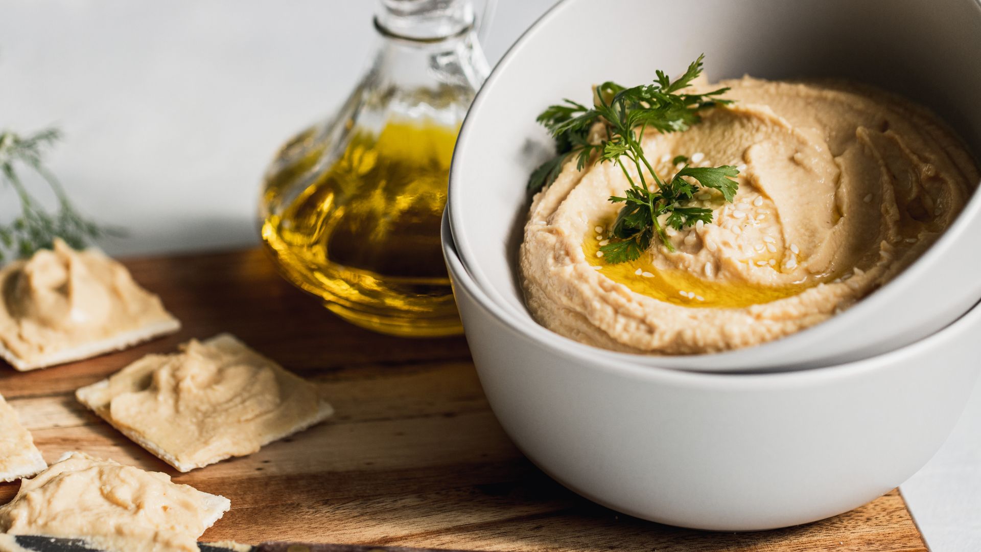A bowl of creamy tahini topped with olive oil and herbs, served with crackers and a bottle of oil in the background.