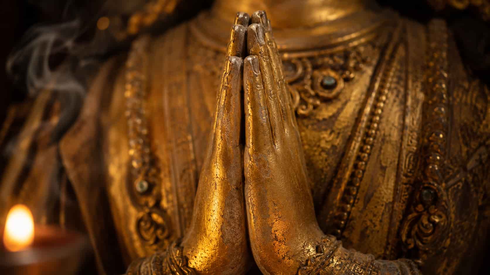 Close-up of a golden Buddha statue’s hands pressed together at the chest in the Anjali Mudra prayer gesture.