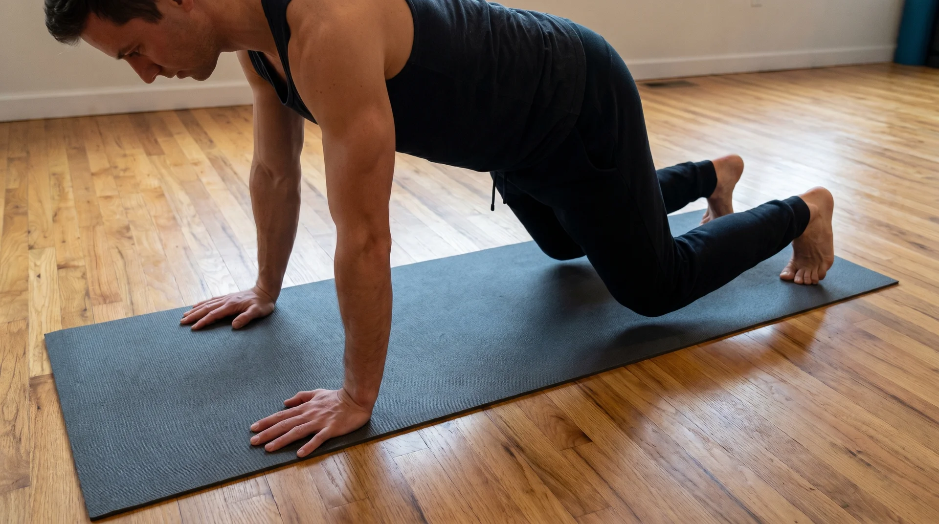 A man in a black tank top and athletic pants performing a bear crawl hold on a dark grey mat over a light-colored wooden floor.