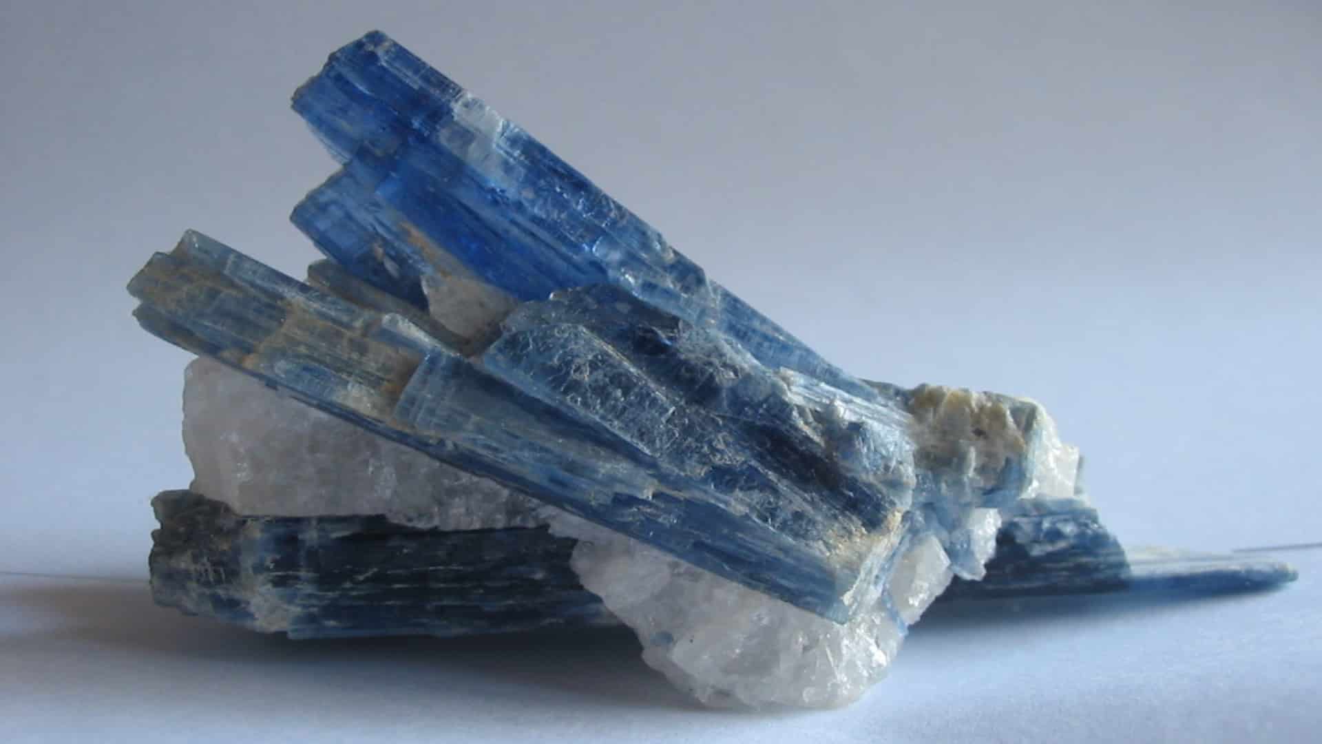 A rough, blade-like blue kyanite crystal with deep blue layers resting horizontally on a white surface.
