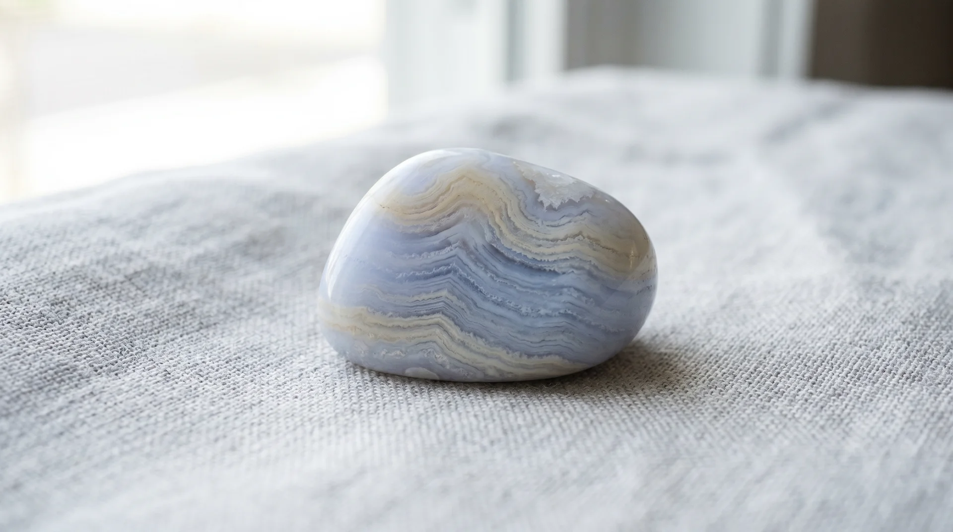 A polished blue lace agate stone with light blue and white bands resting on a light gray fabric surface.