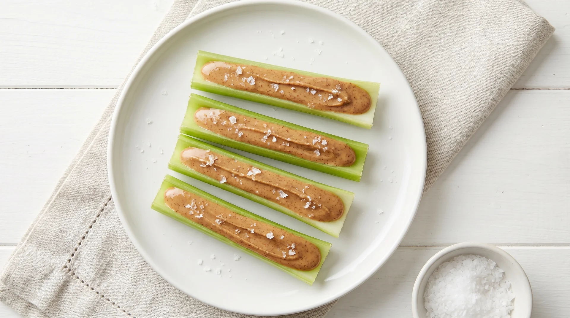 Four celery sticks filled with smooth almond butter, arranged on a white plate with a side bowl of sea salt.