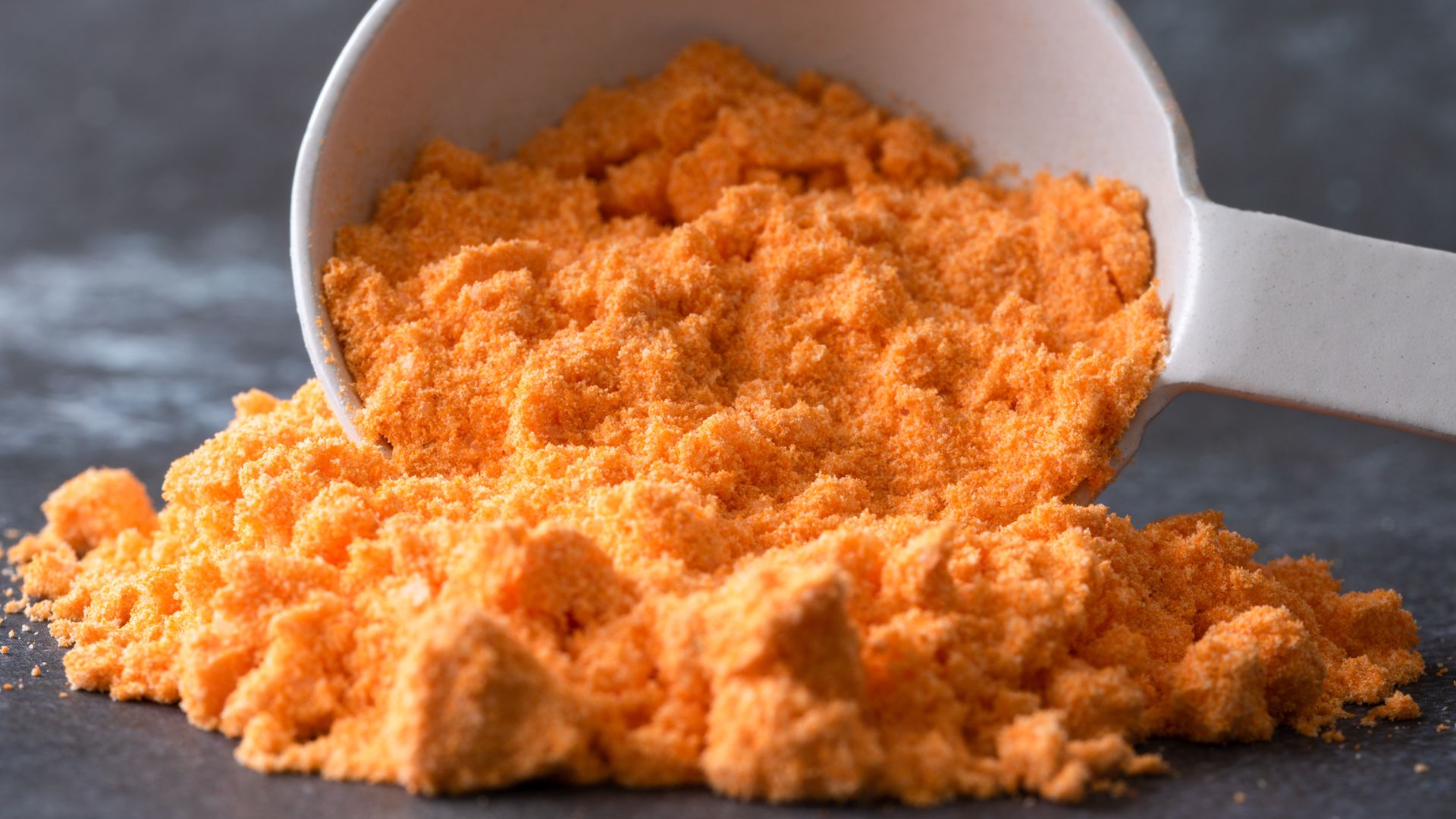 Bright orange cheese powder spilling out of a white measuring scoop onto a textured, dark grey slate background.