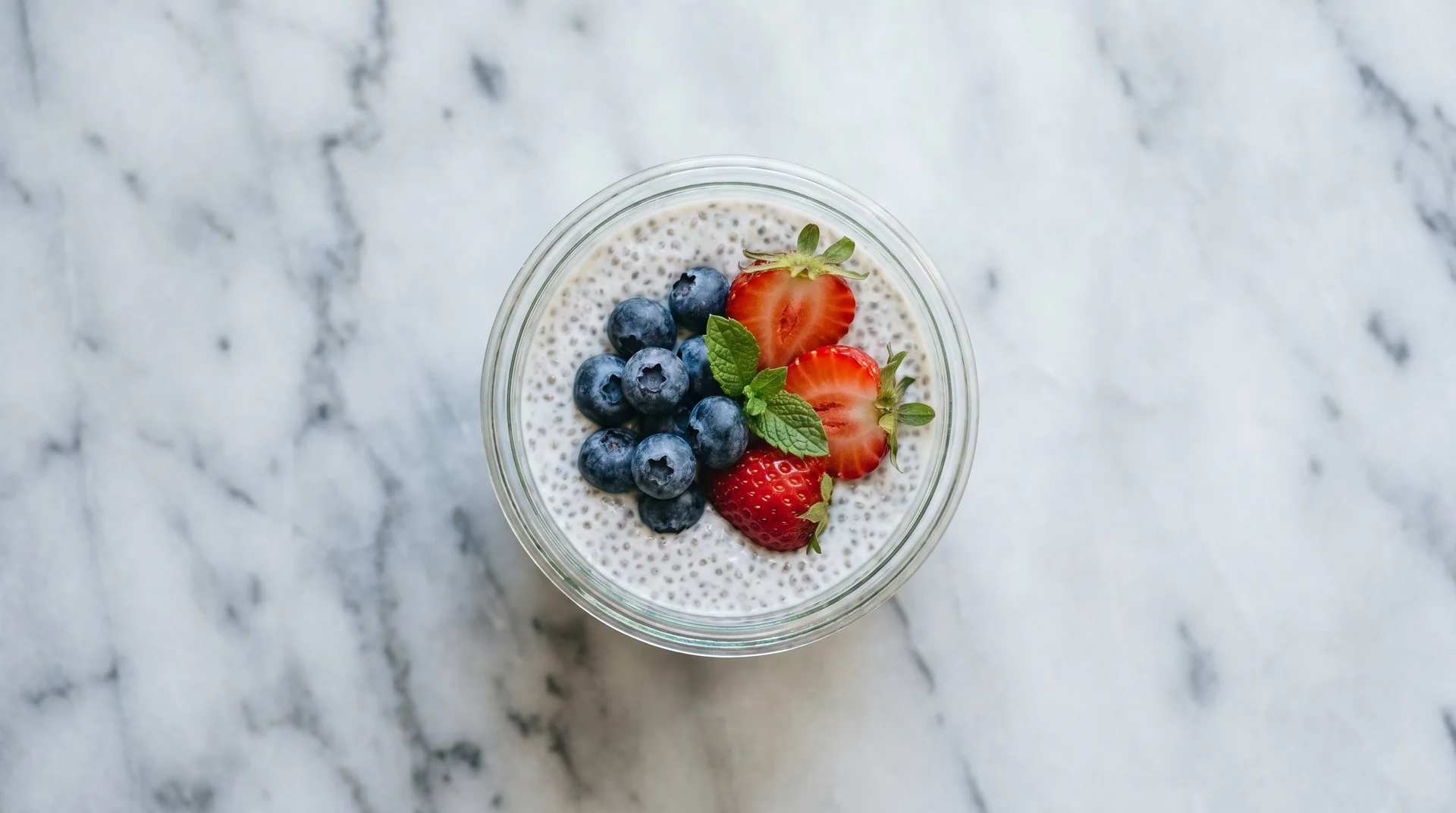 A clear glass bowl of chia seed pudding topped with fresh blueberries, sliced strawberries, and a mint leaf, resting on a white marble surface.