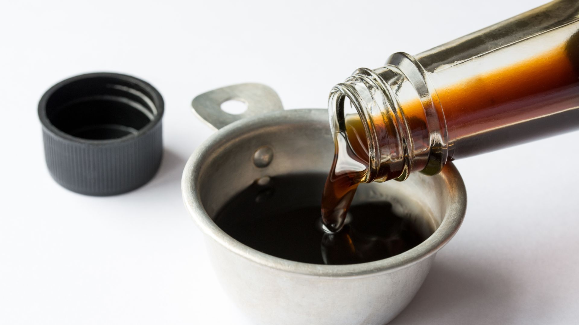 Dark coconut aminos pouring from a clear glass bottle into a small stainless steel measuring cup on a white surface.