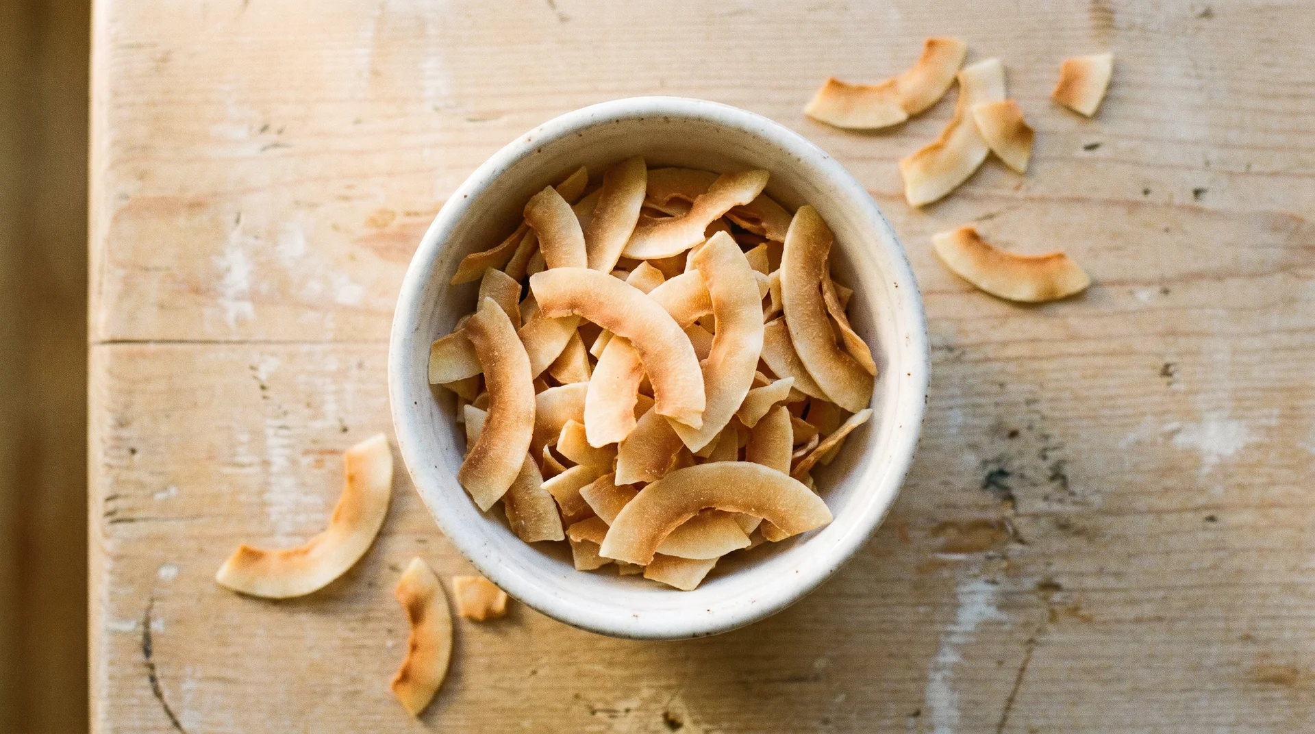 Toasted, golden-brown coconut chips served in a small white ceramic bowl on a rustic wooden table surface.