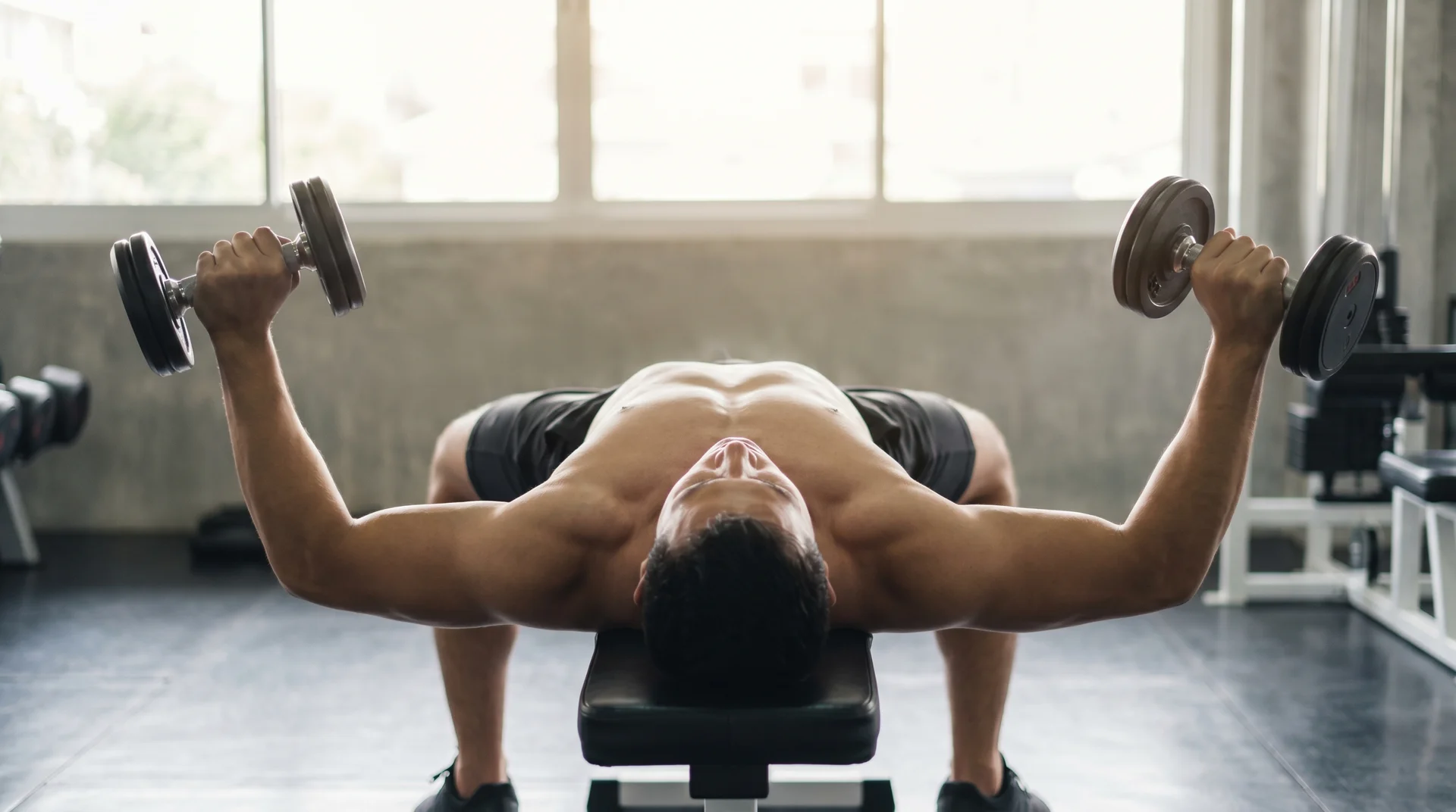 A man lying flat on a gym bench, extending his arms out to the sides while holding a dumbbell in each hand.