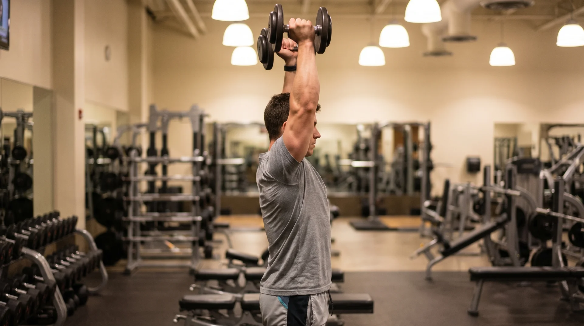 A man standing in a well-equipped gym, pressing a pair of dumbbells straight overhead during a shoulder workout.
