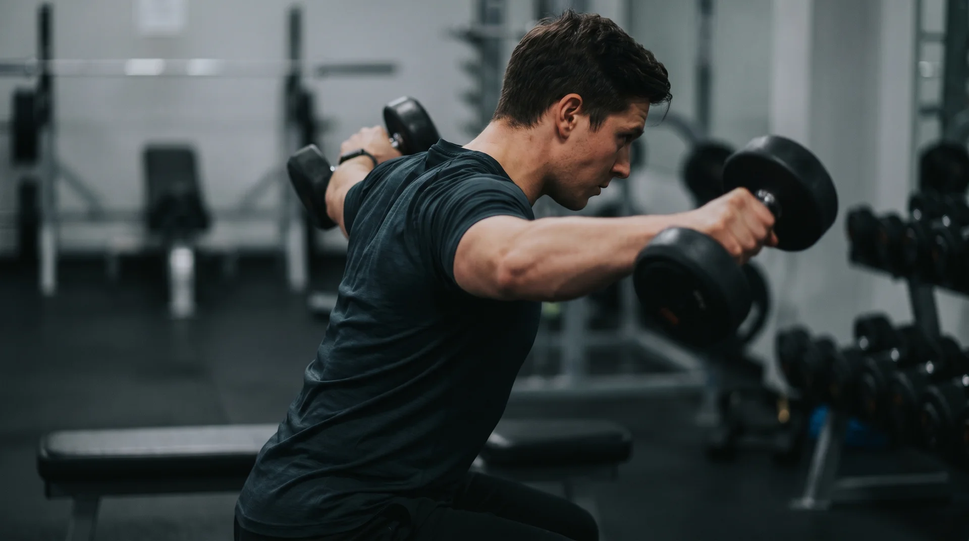 A man sitting on a gym bench, leaning forward and raising dumbbells out to his sides during a rear delt fly exercise.