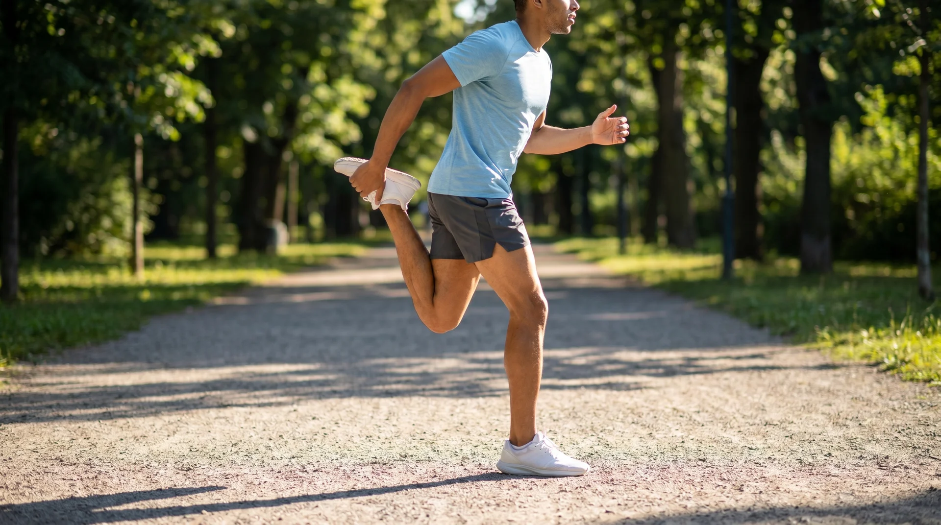 A man jogging outdoors on a paved path, holding his left foot behind his back with his left hand to perform a walking quad stretch.