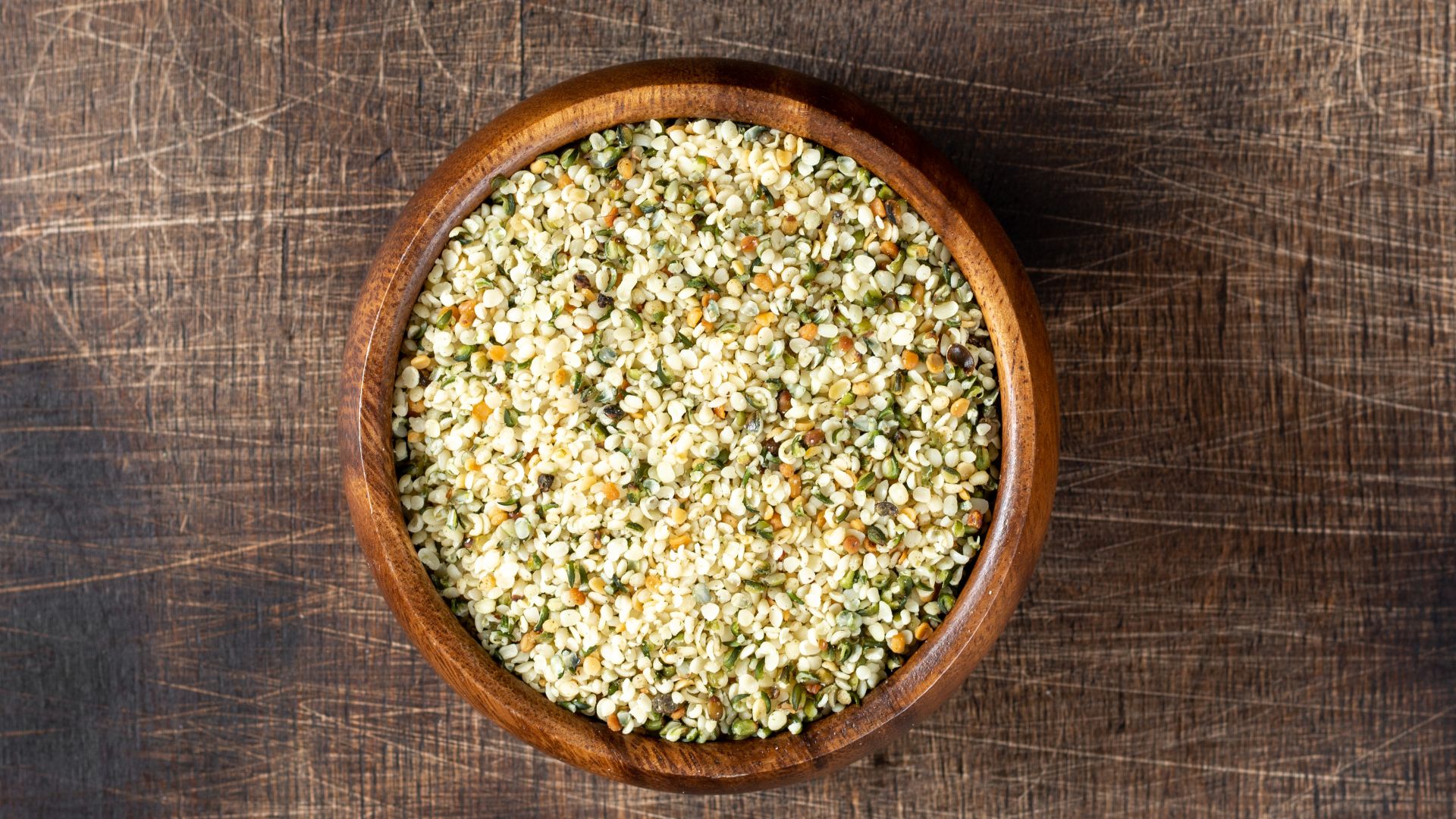 A top-down view of a round wooden bowl filled with shelled hemp seeds, sitting on a dark, textured wood surface.