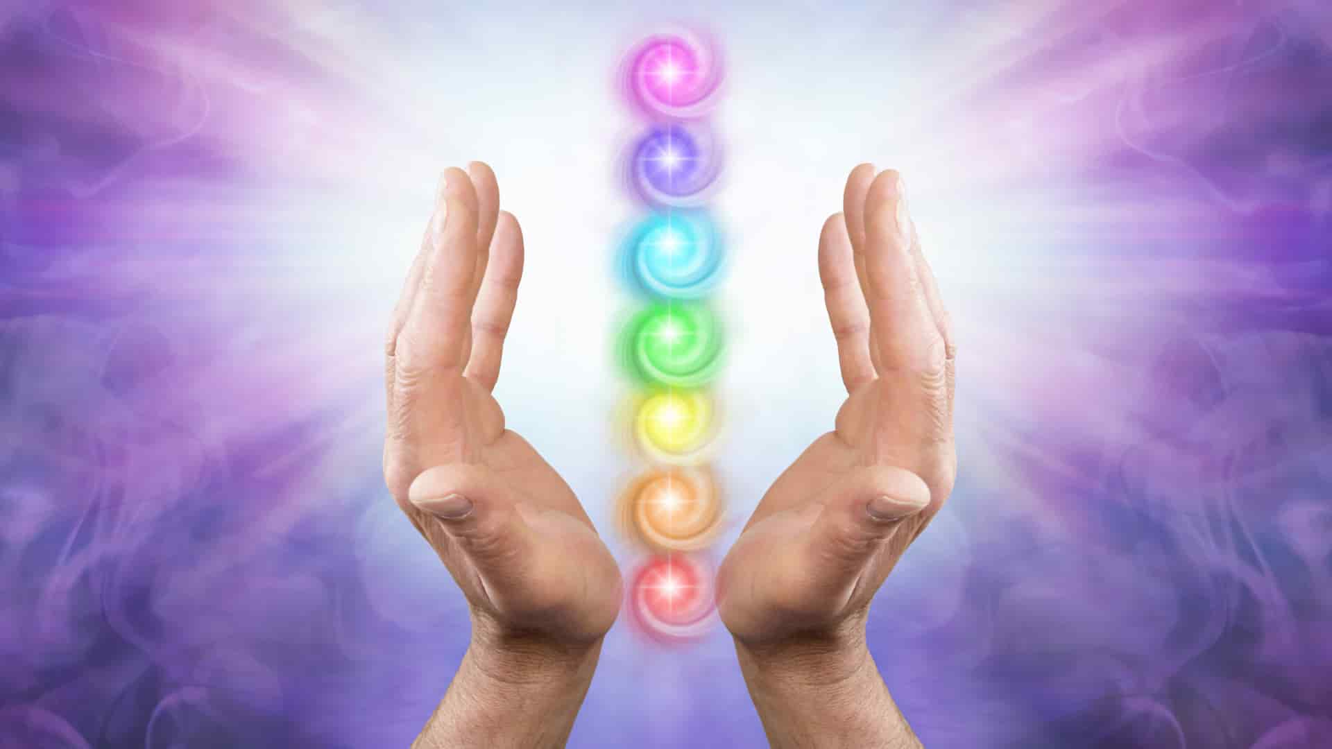 A pair of open hands cupping a vertical alignment of seven glowing, multicolored chakra symbols amidst a soft, swirling purple mist background.