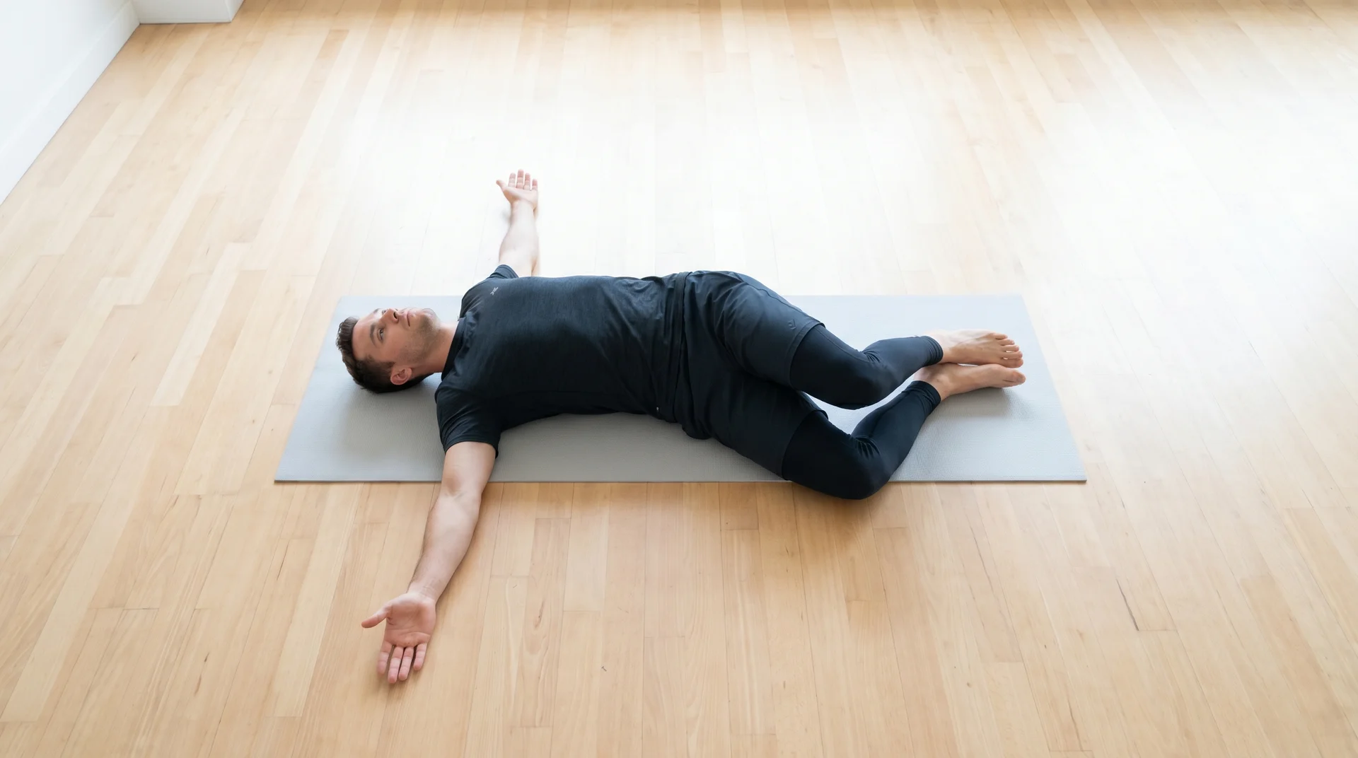 A side-view of a man in athletic wear performing a lying hip rotation stretch on a mat, lowering his bent knees to the side.