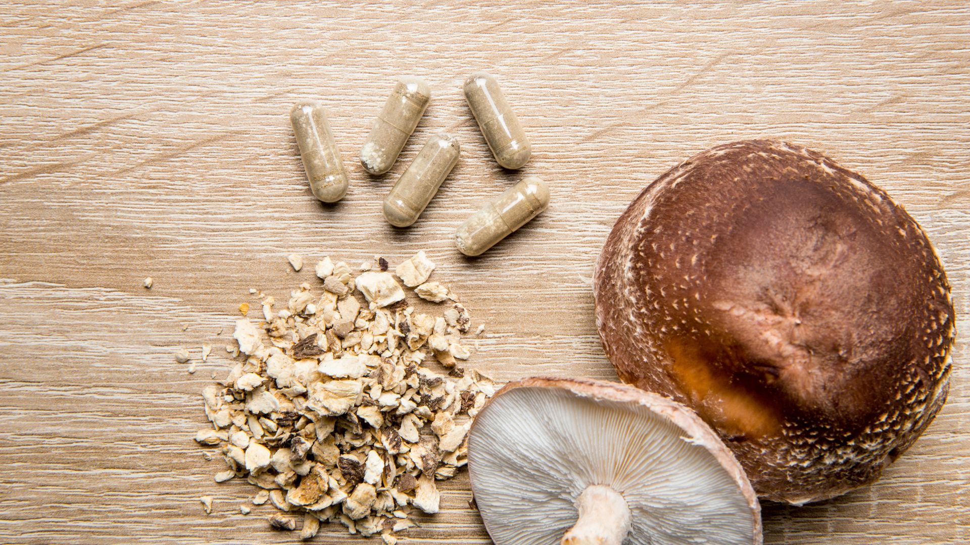 Whole and sliced shiitake mushrooms on a wooden surface next to loose mushroom powder and clear capsule supplements.