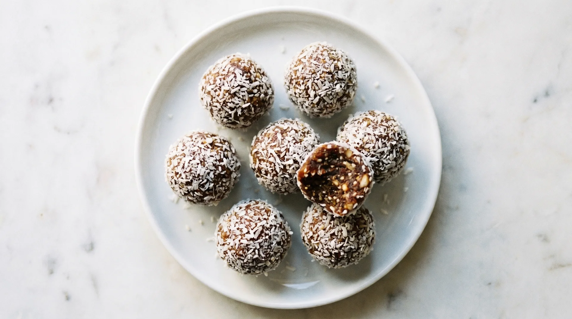 Paleo energy balls coated in shredded coconut on a white ceramic plate with one broken in half to reveal the texture inside.