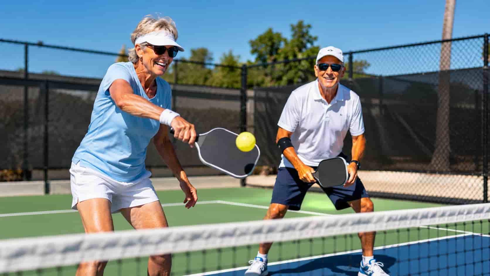An older man and woman smiling and playing pickleball on an outdoor court.