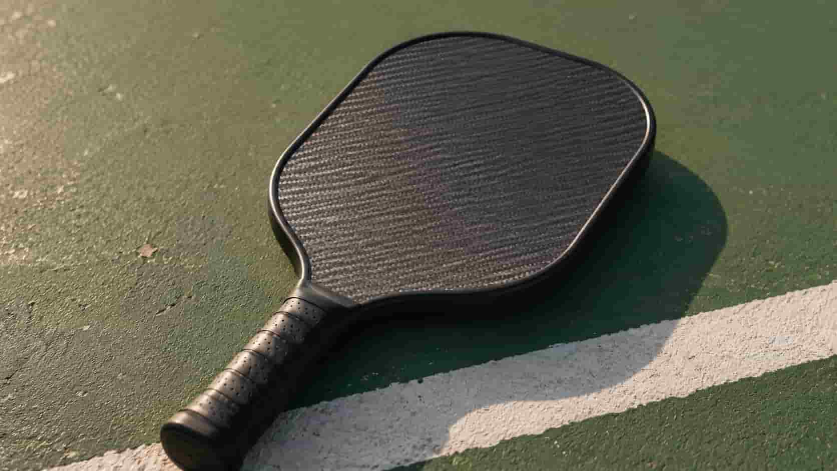 A lightweight black pickleball paddle resting on the surface of an outdoor court.