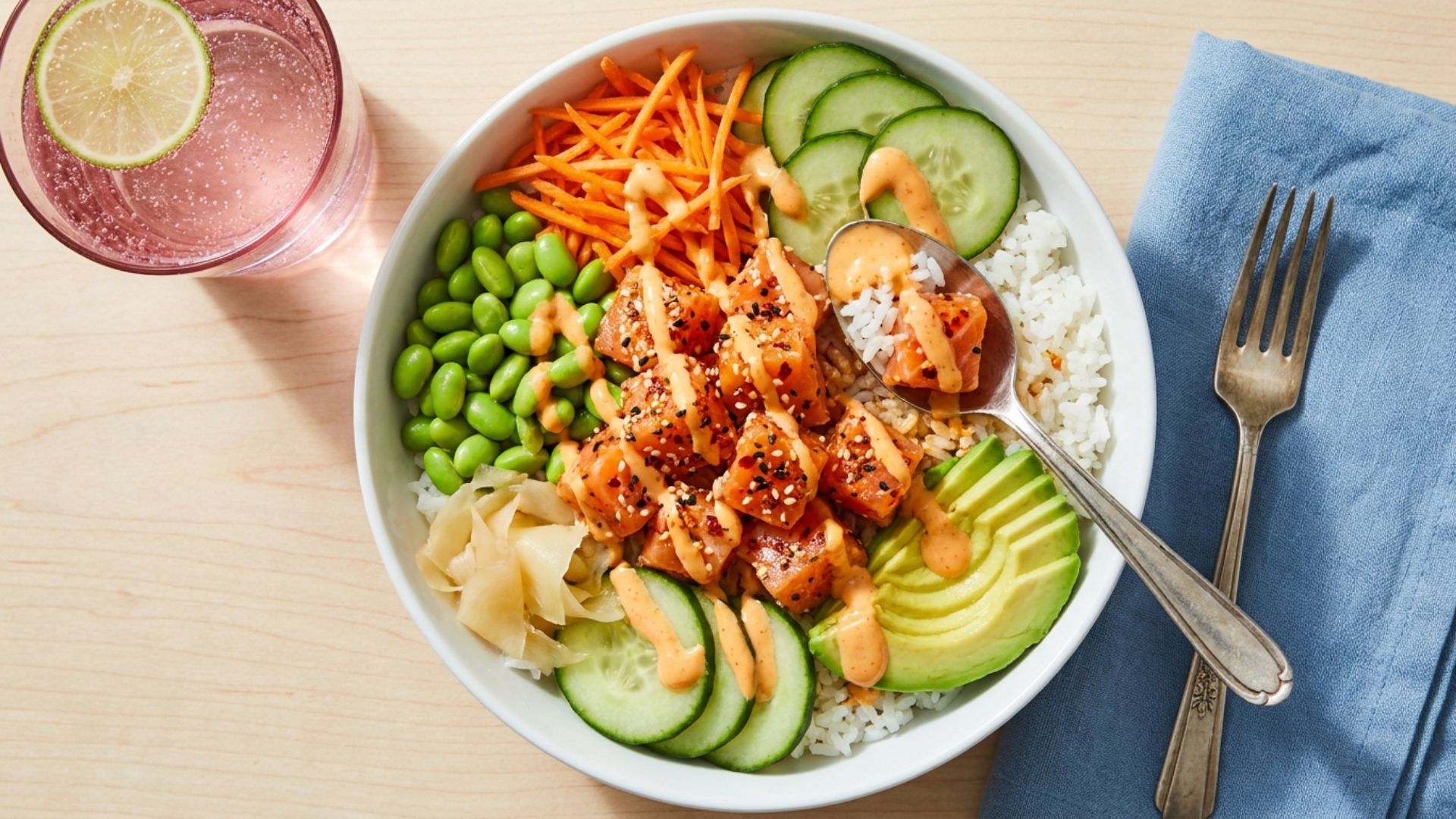 Top-down view of a poke bowl with salmon, rice, avocado, veggies, and a drizzle of spicy mayo next to a lime water.