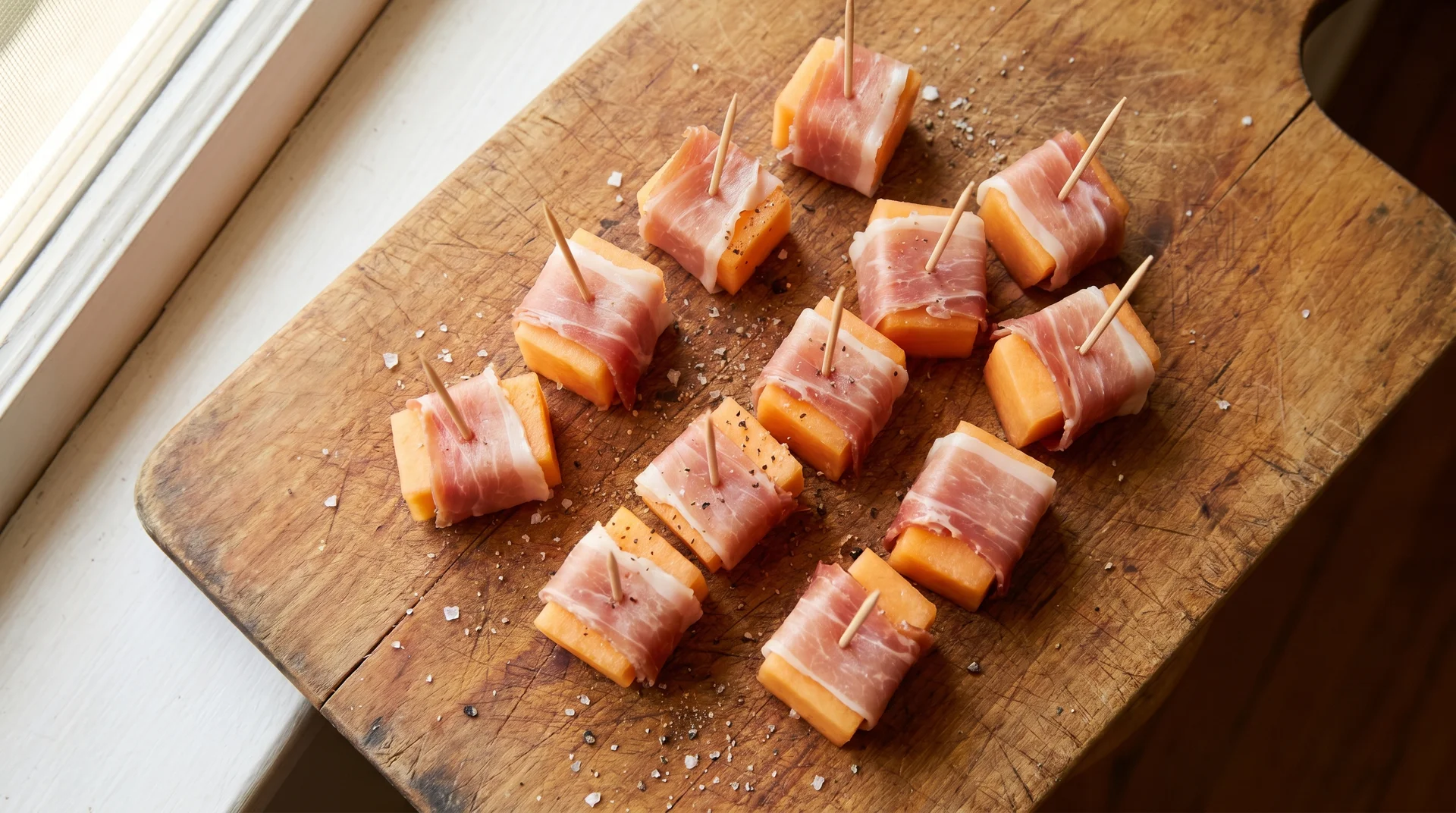 Small cubes of fresh cantaloupe melon wrapped in thin slices of prosciutto, secured with toothpicks on a wooden cutting board.