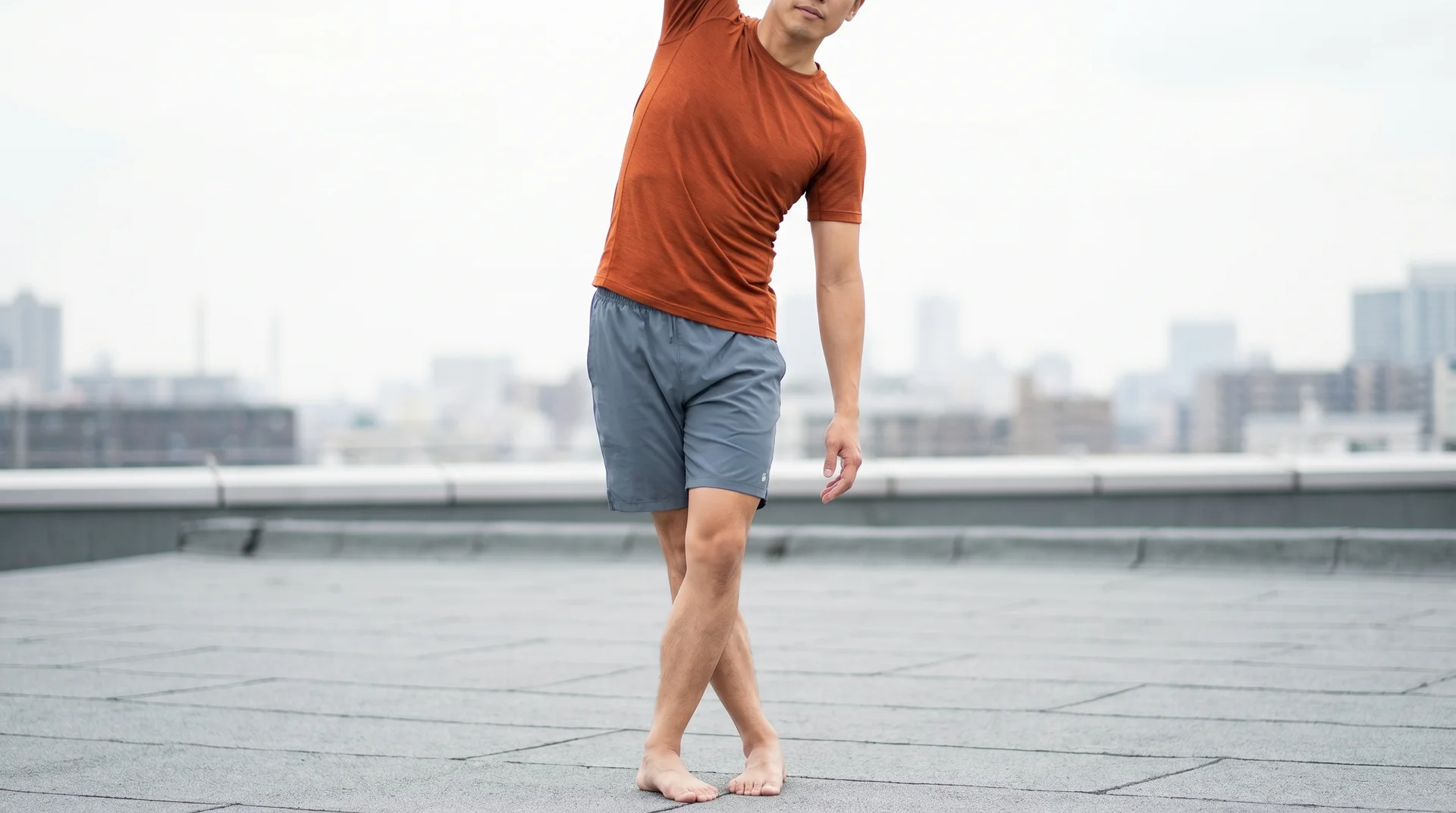 A man standing outdoors, crossing his legs and leaning sideways with one arm overhead to stretch his outer thigh.