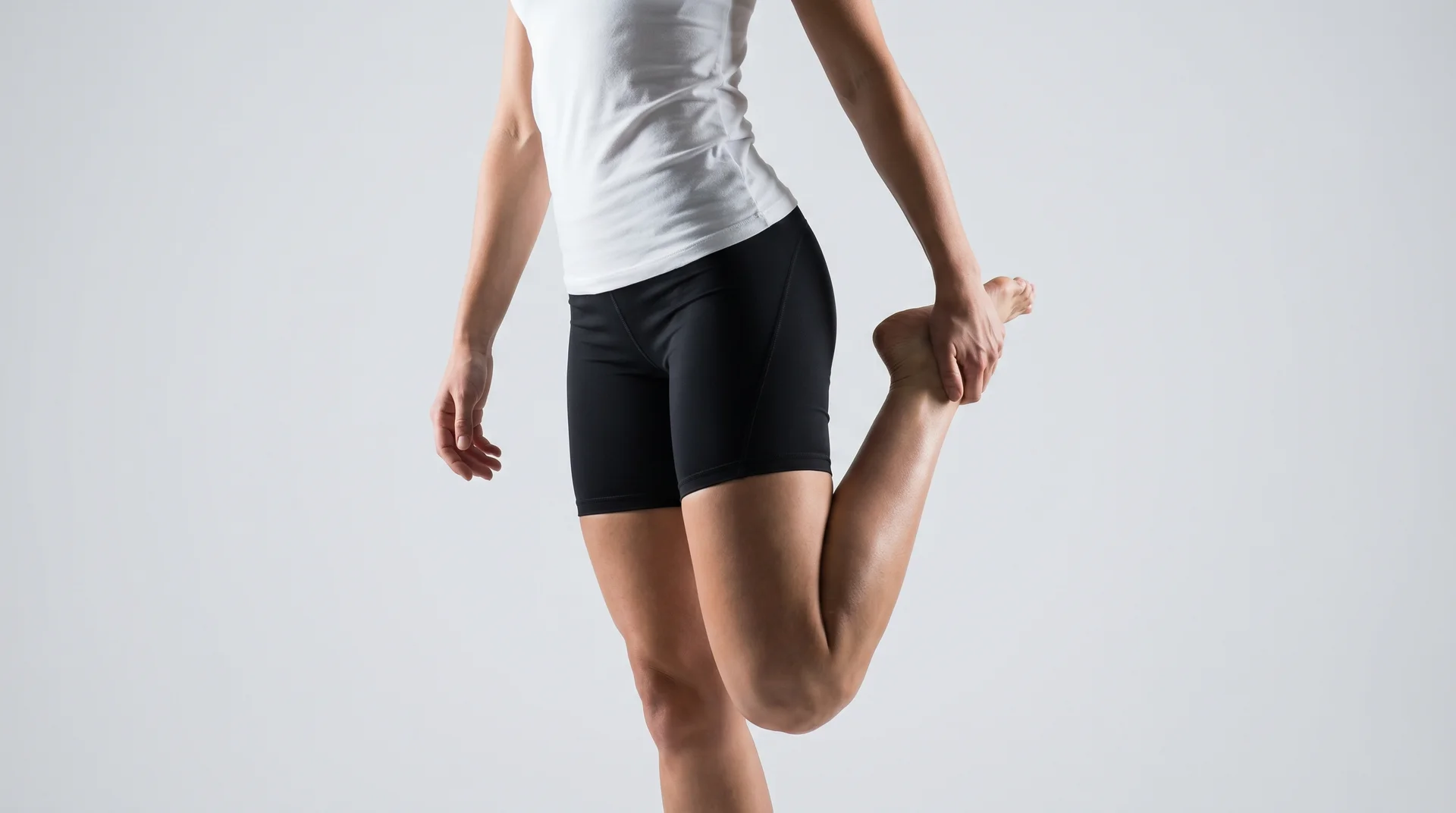 A person wearing a white tank top and black shorts standing indoors, pulling their foot back for a heel-to-glute stretch.
