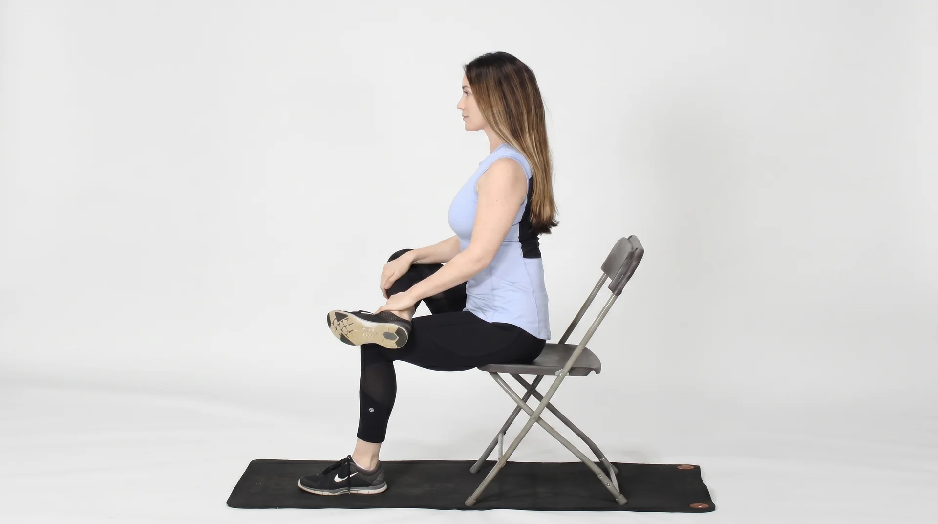 A side-view of a woman in athletic wear sitting upright in a metal folding chair, performing a seated cross-leg hip stretch.