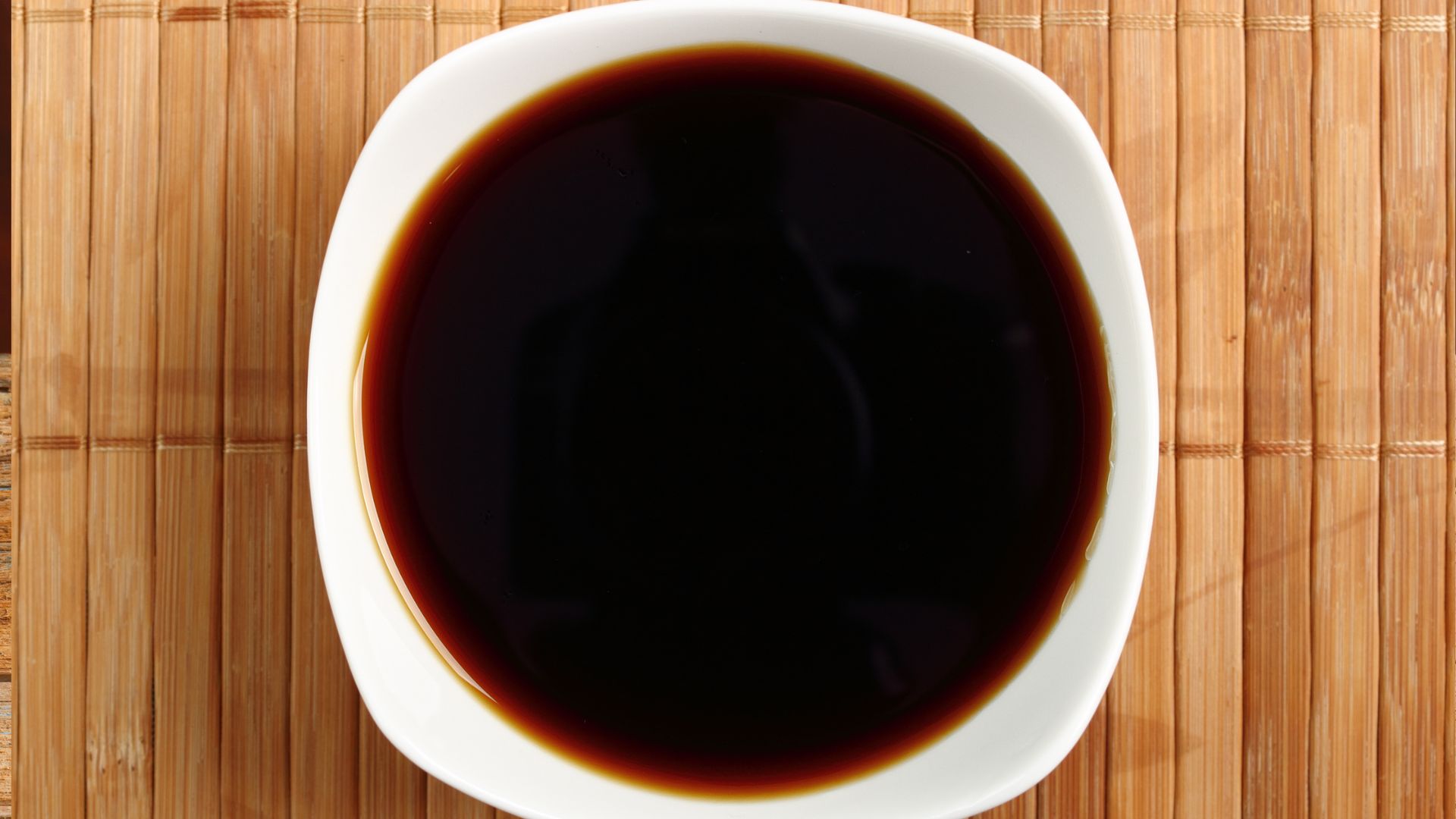 A top-down view of dark soy sauce in a white ceramic bowl, sitting on a natural light-brown bamboo placemat.