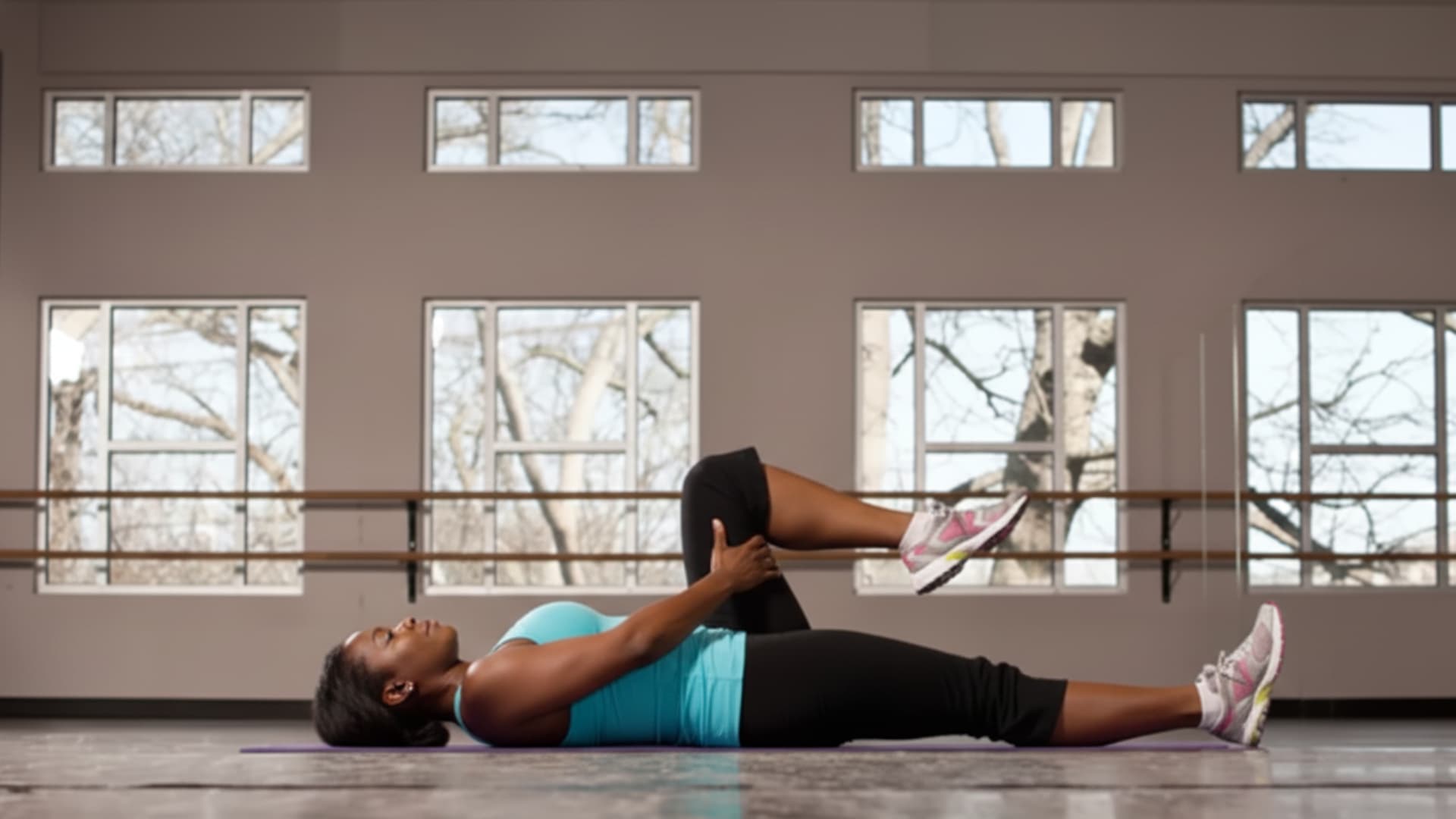 A woman lying on her back on a yoga mat, pulling her right knee toward her chest to perform a supine quad and hip stretch.