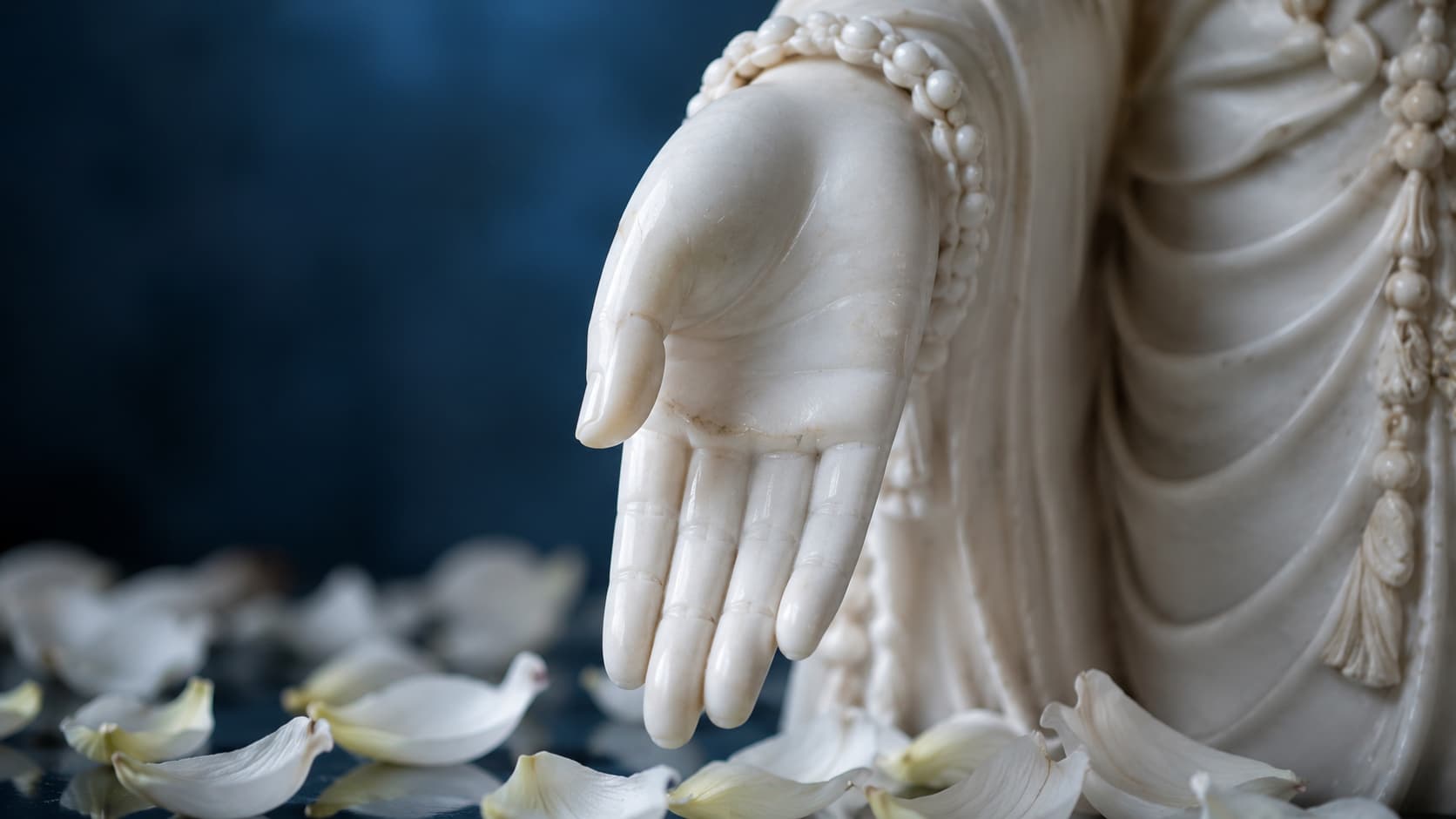 Close-up of a white marble Buddha statue’s hand extended downward in the Varada Mudra gesture, surrounded by white petals.