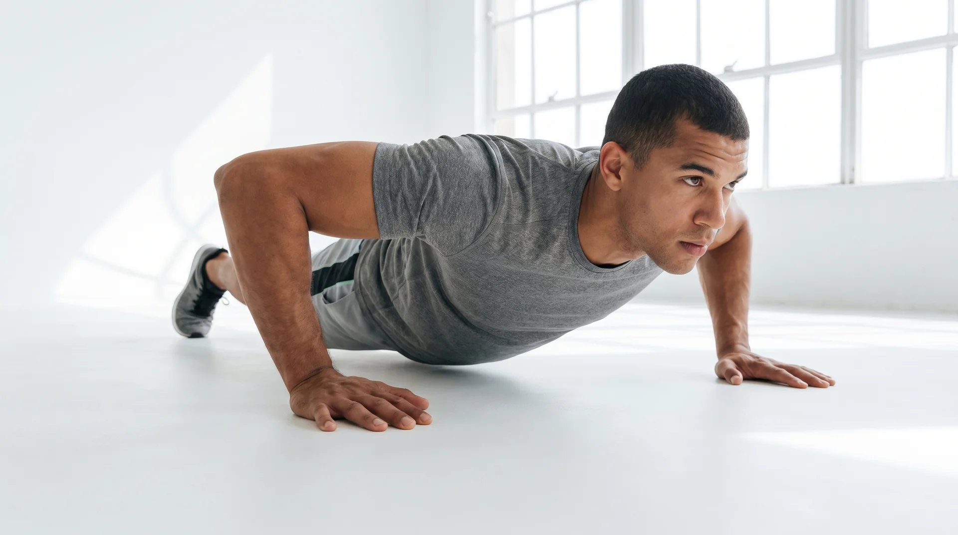 A man with short dark hair performing a wide push-up position on a light-colored floor in a bright studio.